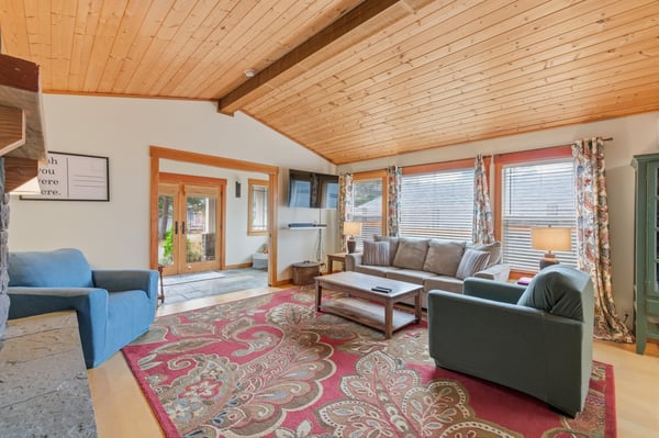Relax in your spacious living room featuring warm wood ceilings, comfortable seating, and large windows that fill the space with natural light.