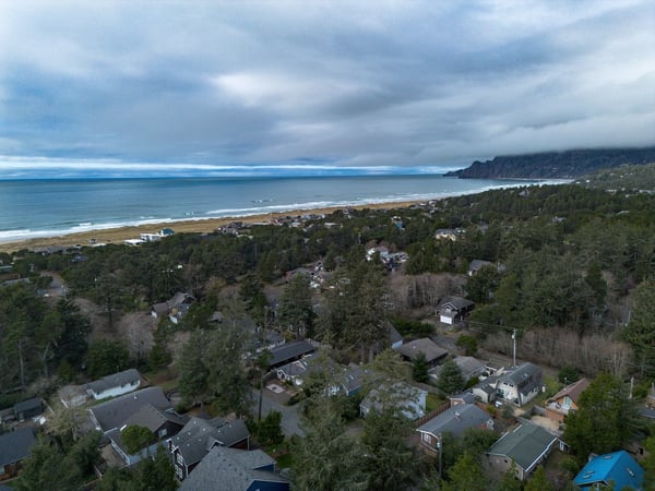 Aerial view of a charming coastal community nestled between lush forest and pristine sandy beach, with dramatic cliffs extending along the shoreline.