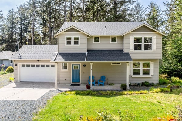 A welcoming two-story home framed by towering evergreens, featuring a covered front porch with cheerful blue chairs and a two-car garage, set on landscaped grounds with a gravel driveway.